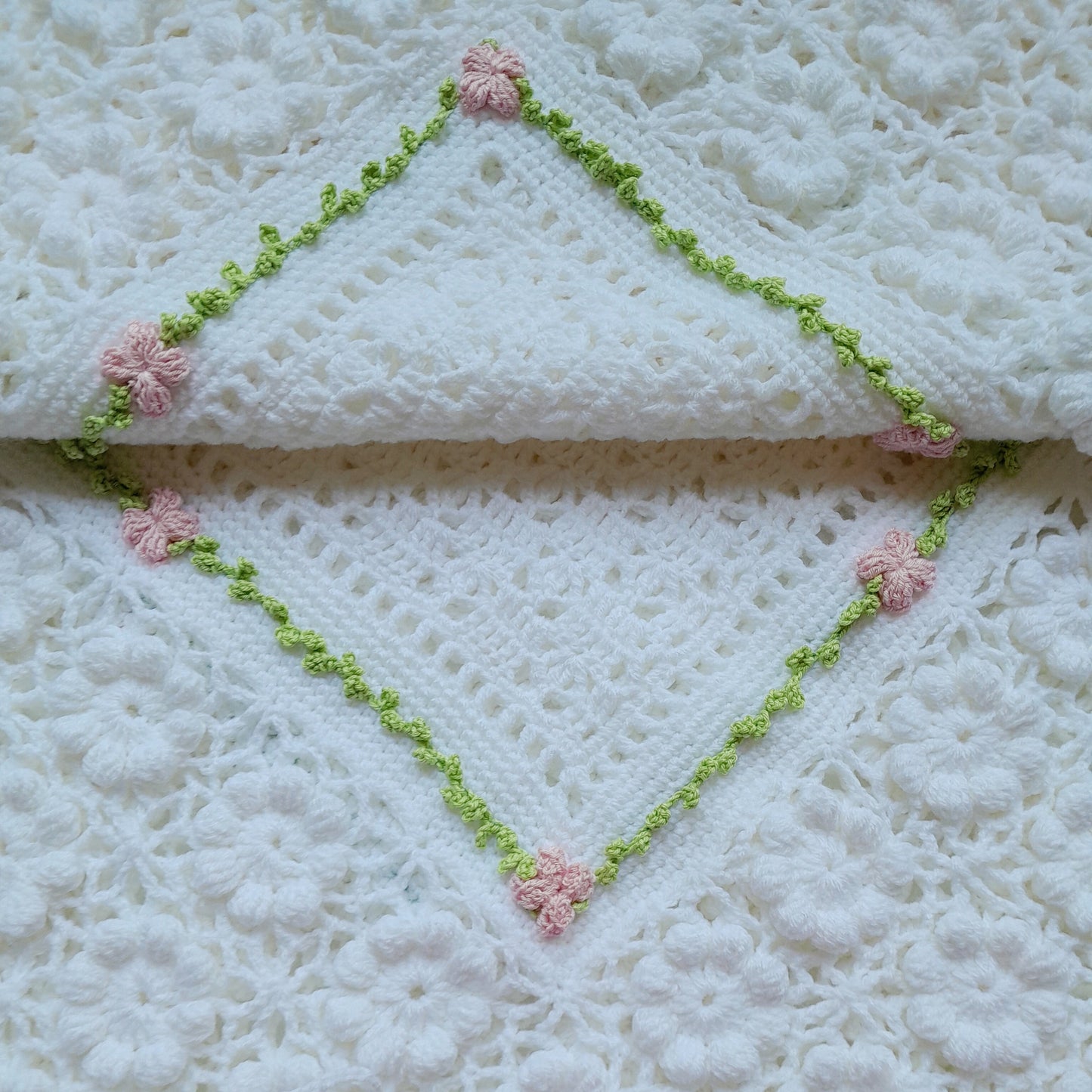 White crocheted blanket with pink flowers and green trim on a white background
