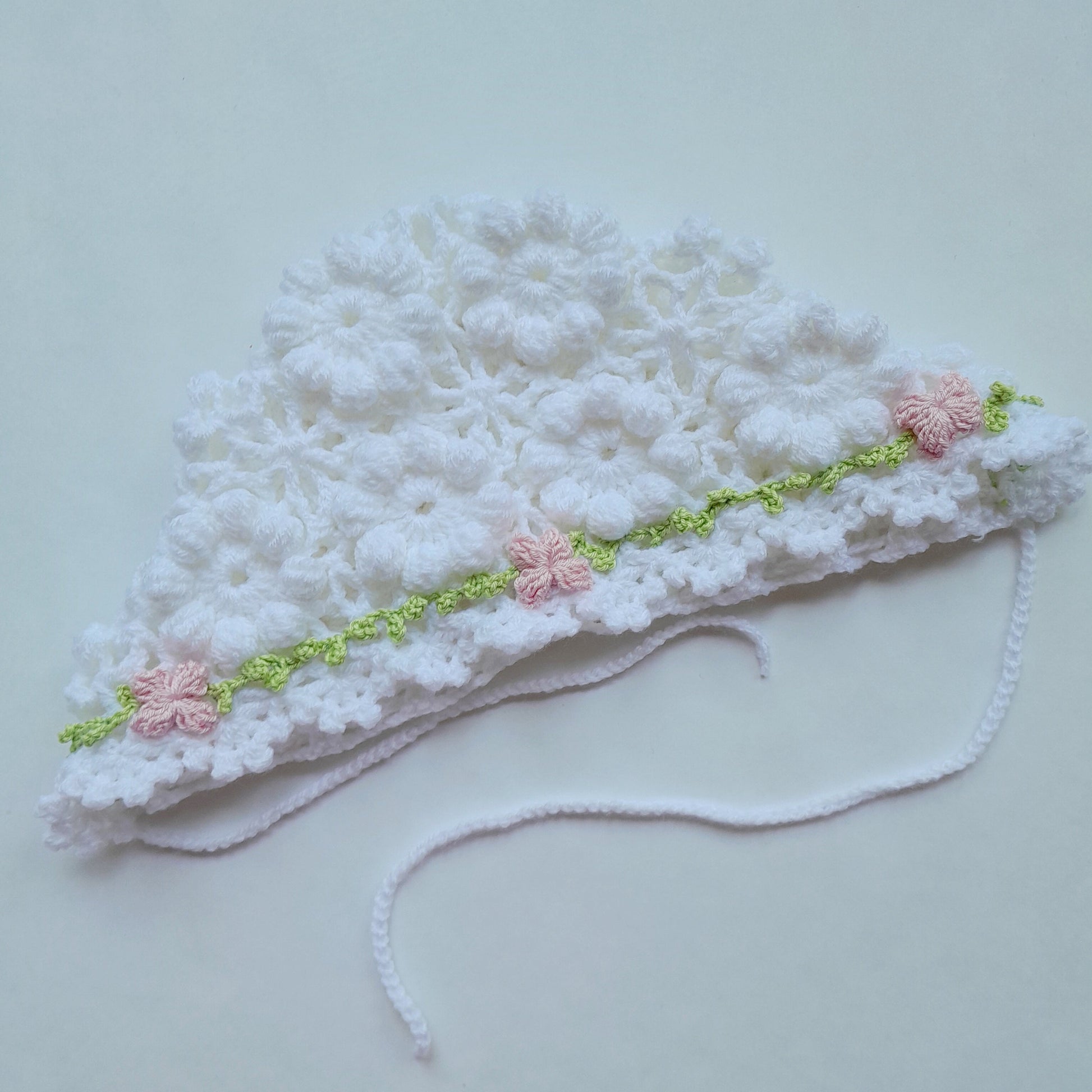 White crocheted baby hat with pink flowers on a light gray background