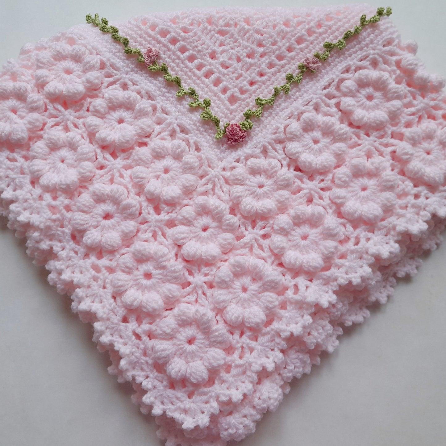 A light pink crocheted baby blanket or shawl with a textured floral pattern and a decorative green and pink beaded border along the pointed top edge, displayed on a light gray background.