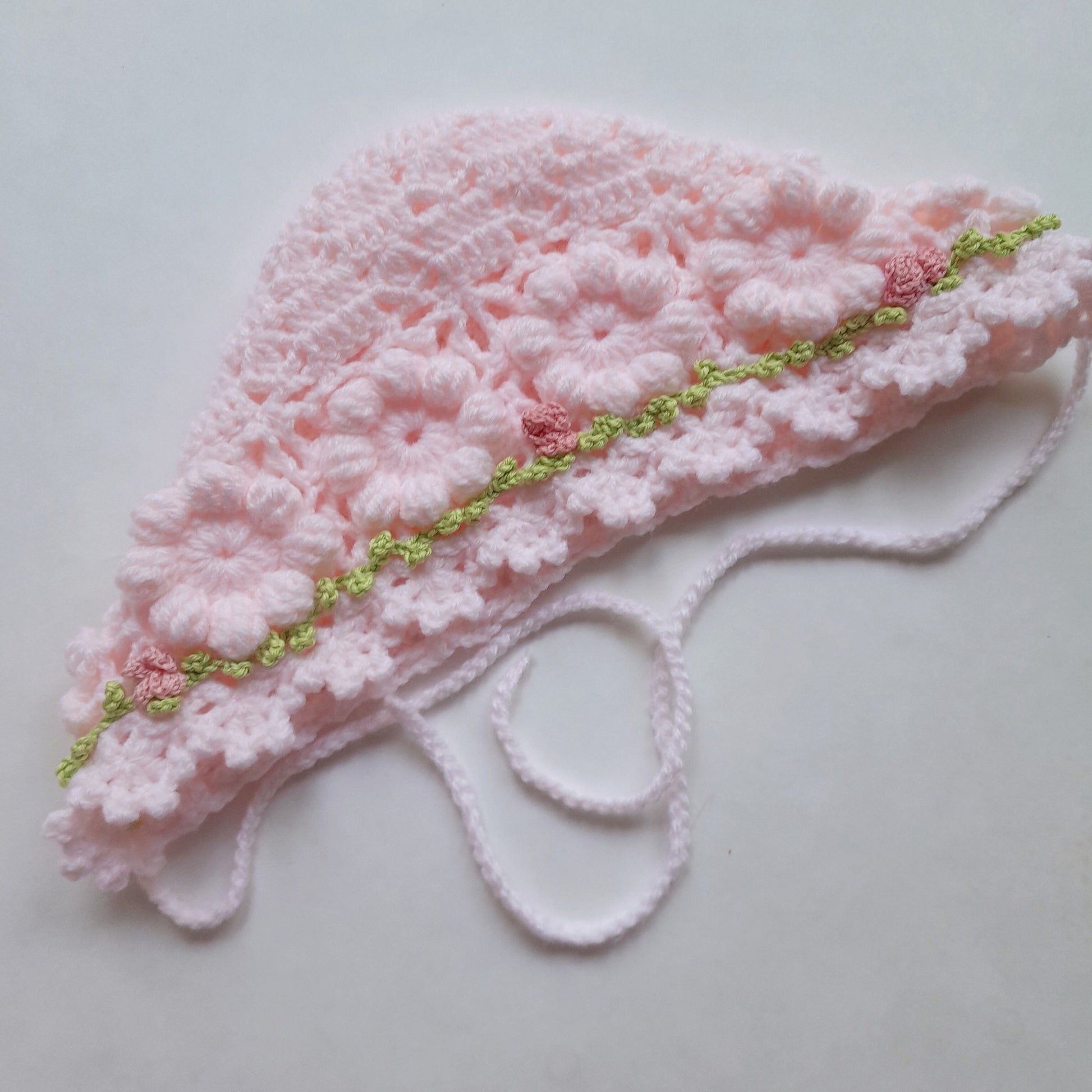 A light pink crocheted bonnet with floral lace detailing and a thin lime green and pink decorative trim, laid flat on a light gray background.