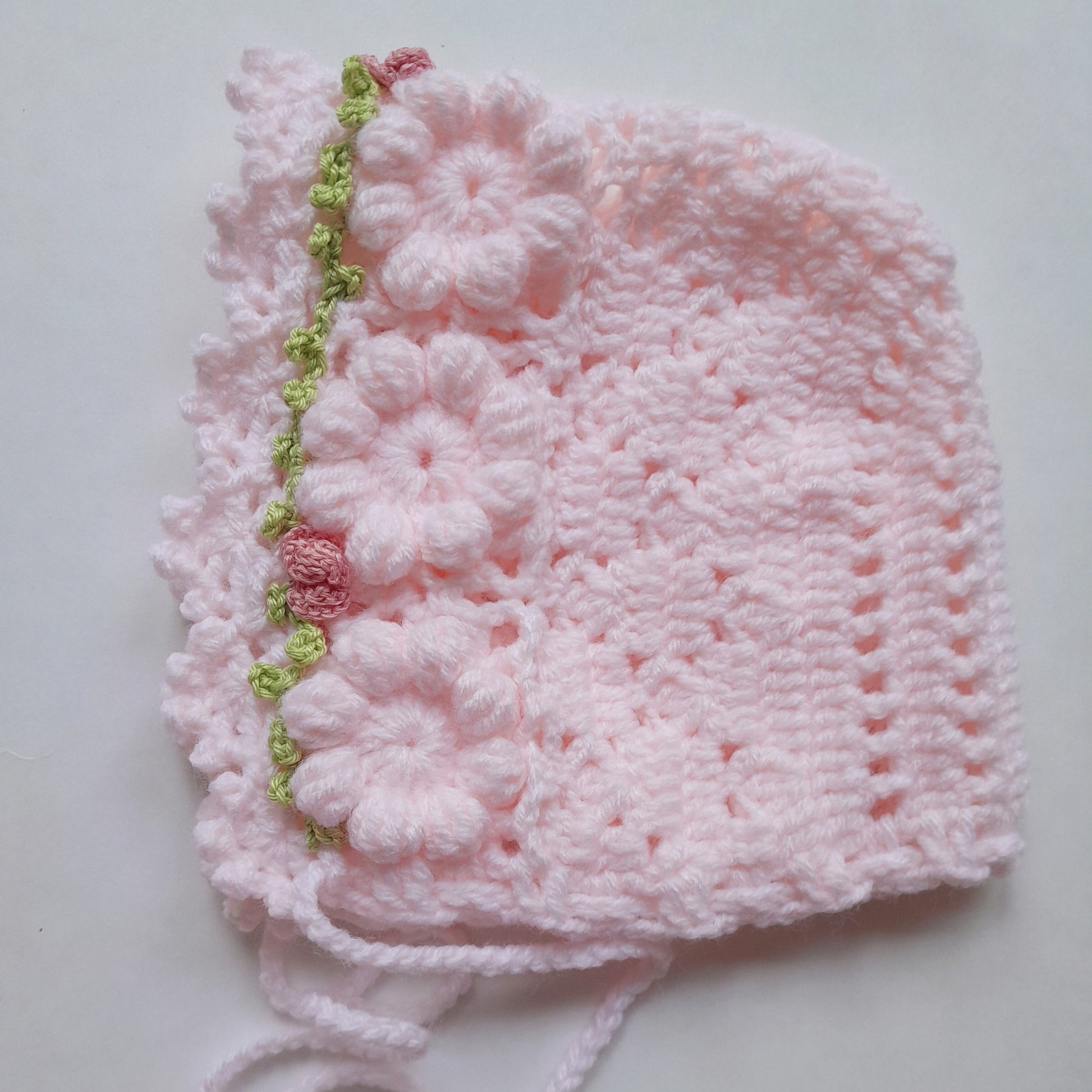A light pink crocheted bonnet with three large floral appliqués and a green and pink decorative trim along the edge, shown on a plain light background.