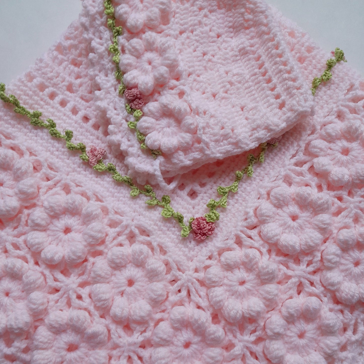 A light pink crocheted baby blanket or shawl with a matching folded corner piece, decorated with a green floral border and small pink flower accents, laid flat on a light background.