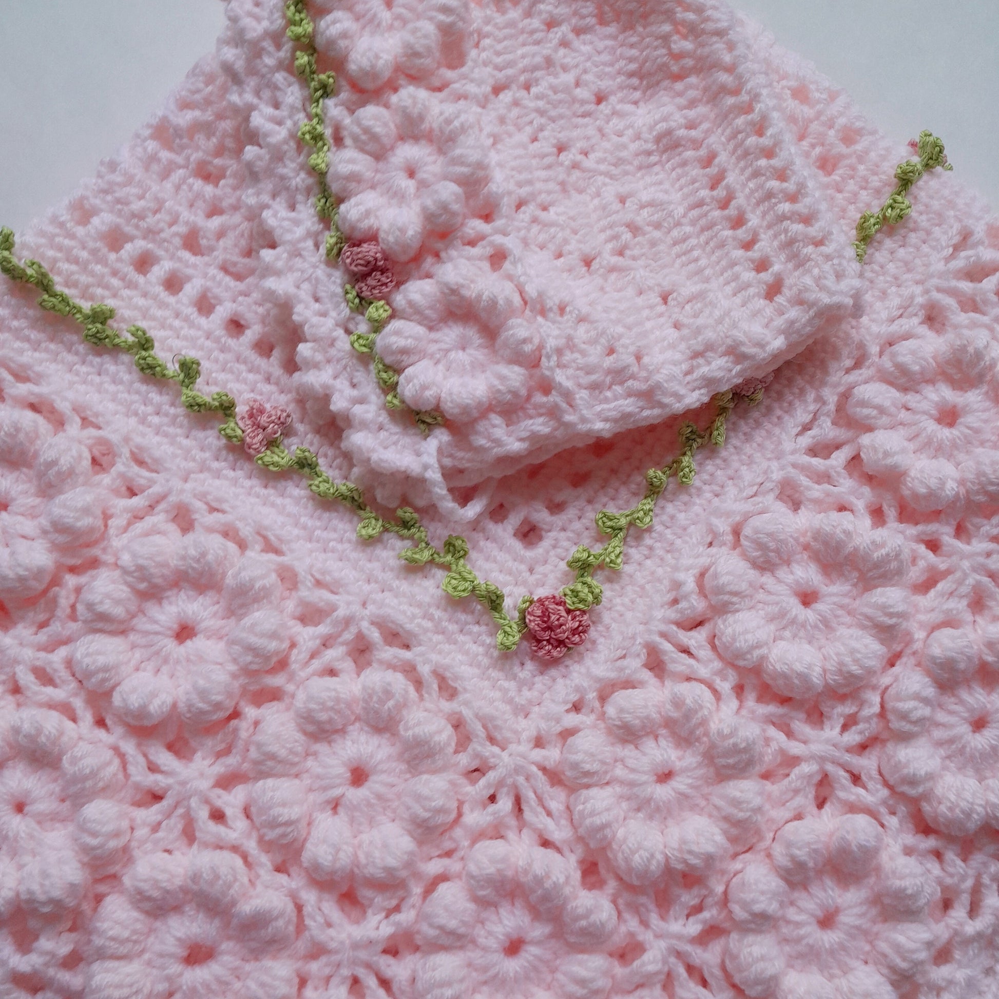 A light pink crocheted baby blanket or shawl with a matching folded corner piece, decorated with a green floral border and small pink flower accents, laid flat on a light background.