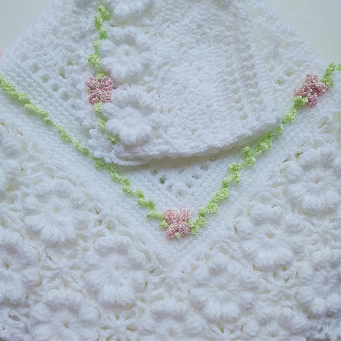 A white crocheted baby blanket or shawl with a matching white bonnet, decorated with a light green floral border and small pink flower accents, laid flat on a light background.