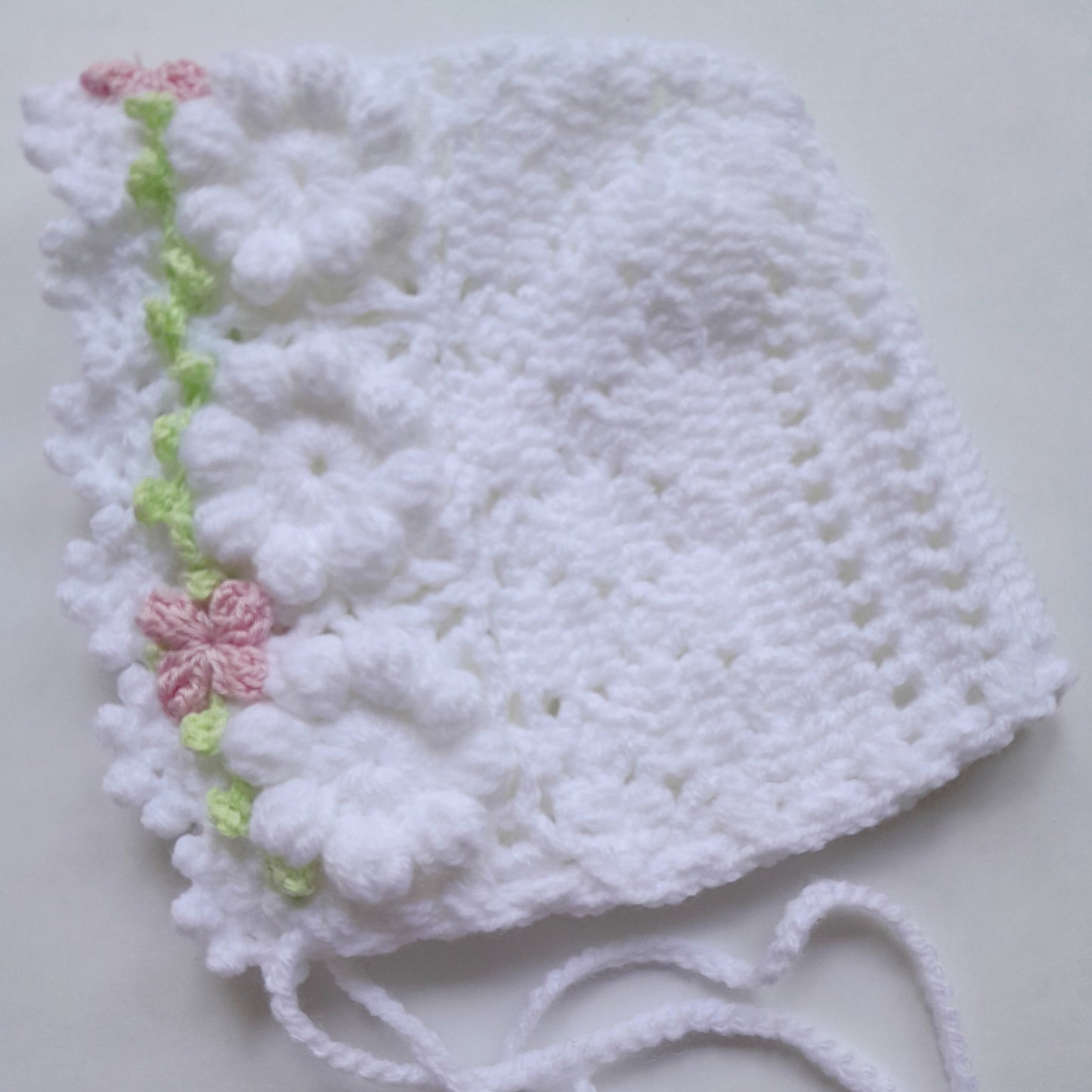 A white crocheted bonnet with a textured openwork pattern, featuring a decorative side border of white daisy-like flowers and two pink and green embroidered flowers, finished with white yarn ties.