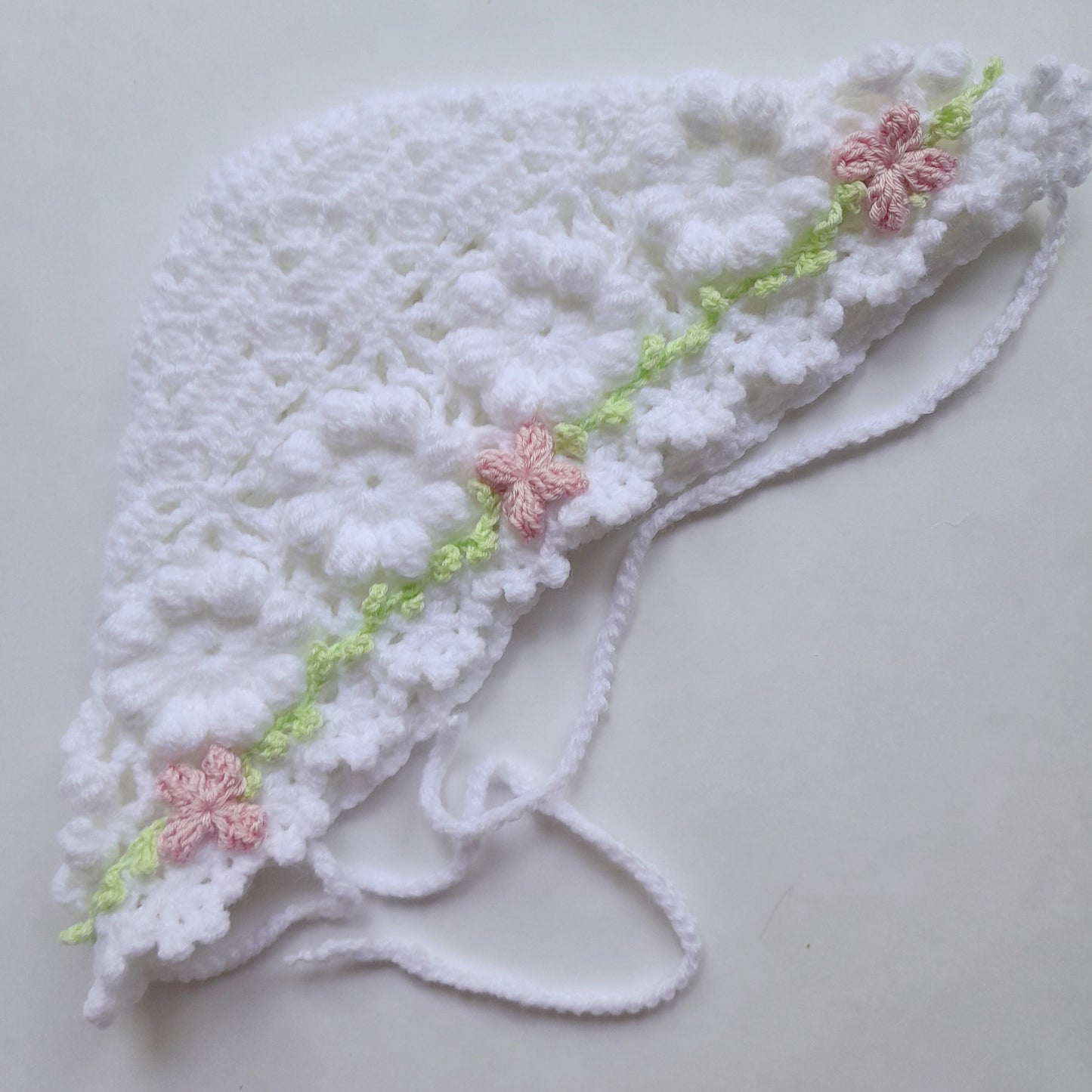 A white crocheted baby bonnet with delicate pink floral accents and a light green embroidered vine detail, laid flat on a light gray background.