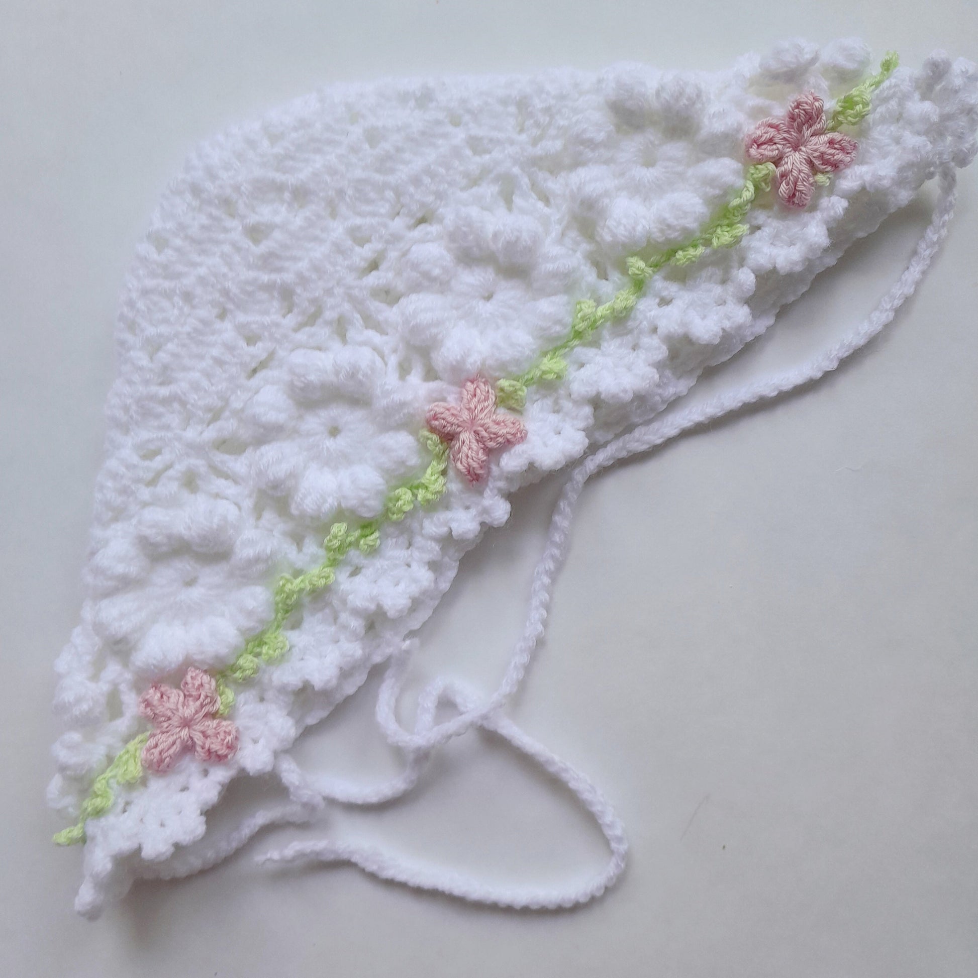 A white crocheted baby bonnet with delicate pink floral accents and a light green embroidered vine detail, laid flat on a light gray background.
